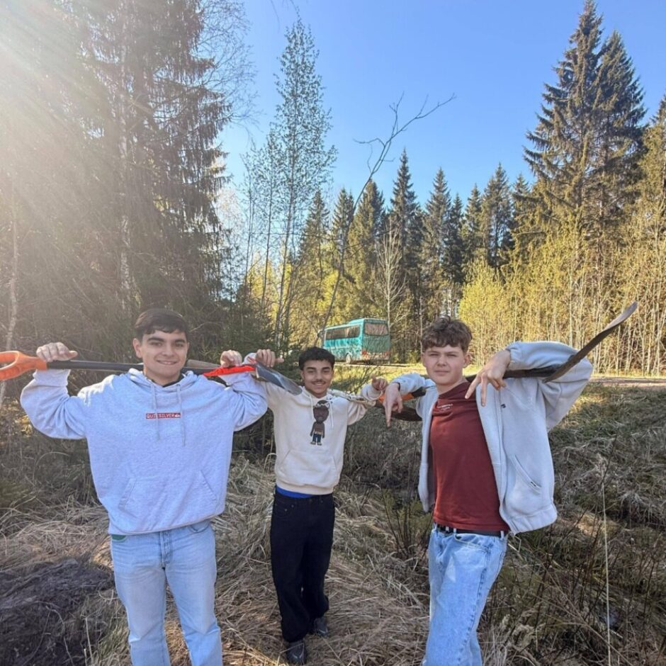 Trīs smaidīgi pusaudži stāv ārā, turot lāpstas uz pleciem. Viņi atrodas saulainā, zālainā vietā ar mežu fonā, kur redzams arī gaiši zils transportlīdzeklis.