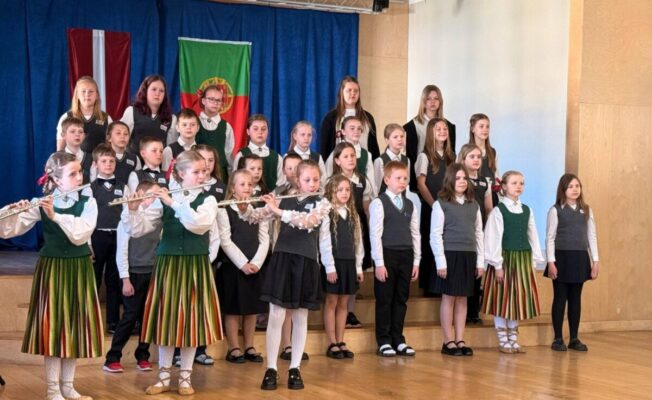 Grupa bērnu uzstājas uz koka skatuves. Priekšplānā trīs meitenes latviešu tautastērpos spēlē flautas. Aiz viņām rindās stāv citi bērni, daži skolas formās, daži tautastērpos, iespējams, kā koris. Fonā uz zila aizkara redzami Latvijas un Portugāles karogi.