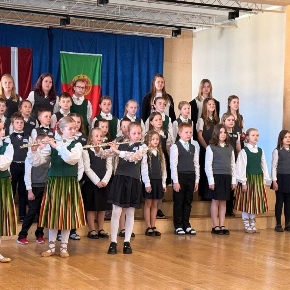 Grupa bērnu uzstājas uz koka skatuves. Priekšplānā trīs meitenes latviešu tautastērpos spēlē flautas. Aiz viņām rindās stāv citi bērni, daži skolas formās, daži tautastērpos, iespējams, kā koris. Fonā uz zila aizkara redzami Latvijas un Portugāles karogi.