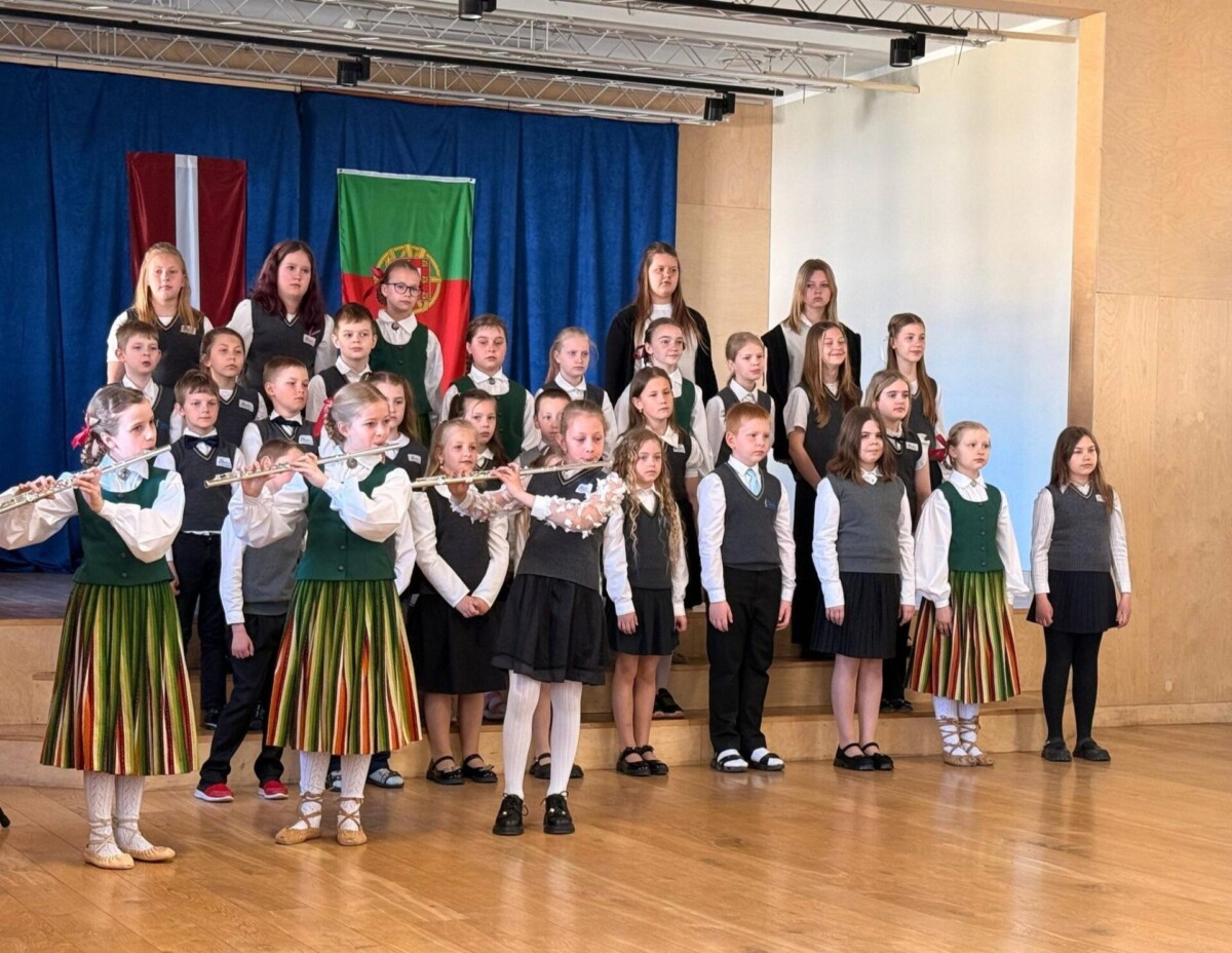 Grupa bērnu uzstājas uz koka skatuves. Priekšplānā trīs meitenes latviešu tautastērpos spēlē flautas. Aiz viņām rindās stāv citi bērni, daži skolas formās, daži tautastērpos, iespējams, kā koris. Fonā uz zila aizkara redzami Latvijas un Portugāles karogi.