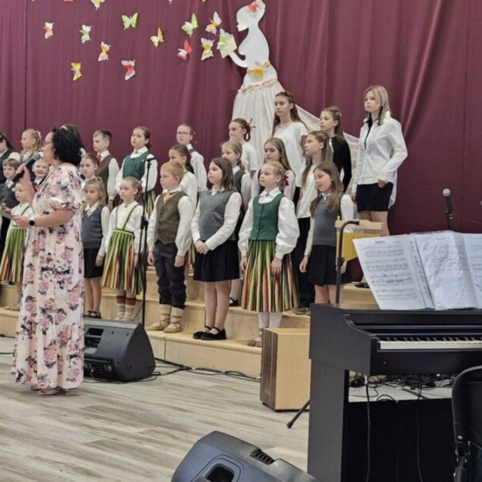Sieviete ziedu kleitā runā mikrofonā, vadot bērnu kori, kas uz skatuves tērpts stilizētos tautas tērpos. Fonā ir sarkans aizkars ar sievietes siluetu un krāsainiem tauriņiem. Pa labi redzama sieviete spēlē klavieres.