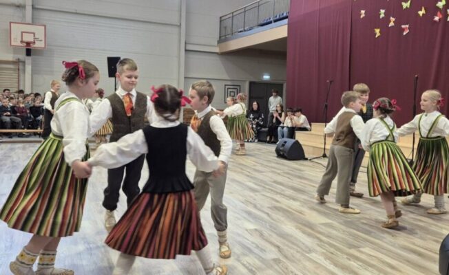 Bērni latviešu tautas tērpos dejo aplī sporta zālē, skatītāju priekšā. Zāles gala sienu rotā sarkani aizkari ar papīra tauriņiem un balta sievietes figūra.