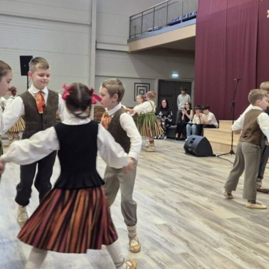 Bērni latviešu tautas tērpos dejo aplī sporta zālē, skatītāju priekšā. Zāles gala sienu rotā sarkani aizkari ar papīra tauriņiem un balta sievietes figūra.