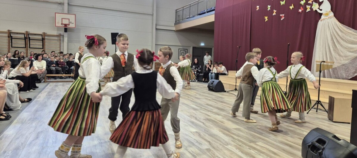 Bērni latviešu tautas tērpos dejo aplī sporta zālē, skatītāju priekšā. Zāles gala sienu rotā sarkani aizkari ar papīra tauriņiem un balta sievietes figūra.