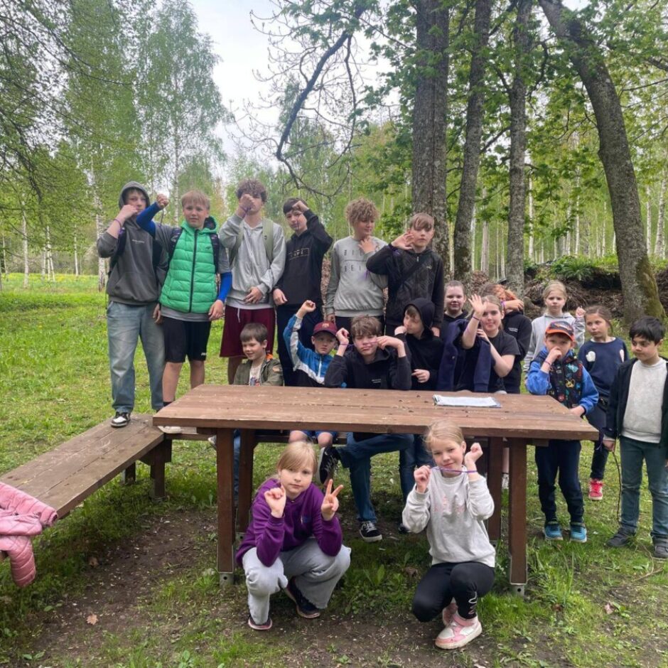 Bērnu un pusaudžu grupa pozē pie koka piknika galda brīvā dabā, mežainā pļavā. Daži no viņiem smaida, citi rāda rotaļīgus žestus.