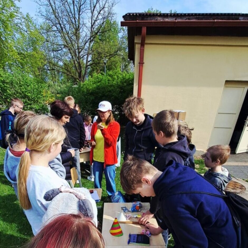 Bērni un pieaugušie pulcējas ap koka galdiem ārā. Zēni un meitenes darbojas ar priekšmetiem uz galda, tostarp krāsainu svītrainu konusu. Sieviete sarkanā jakā un baltā cepurē sarunājas ar bērniem. Aina norisinās saulainā dienā uz zaļas zāles ar kokiem un ēku fonā.