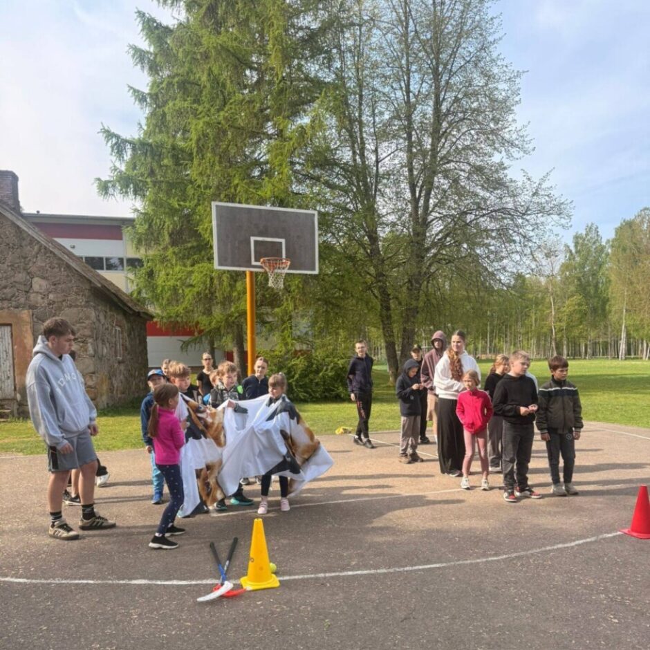 Grupa bērnu un pieaugušo piedalās āra aktivitātē uz basketbola laukuma. Daži bērni tur lielu auduma palagu ar suņa attēlu, kamēr citi stāv apkārt. Uz asfalta ir izvietoti sporta konusi, tostarp dzeltens konuss ar uzrakstu 