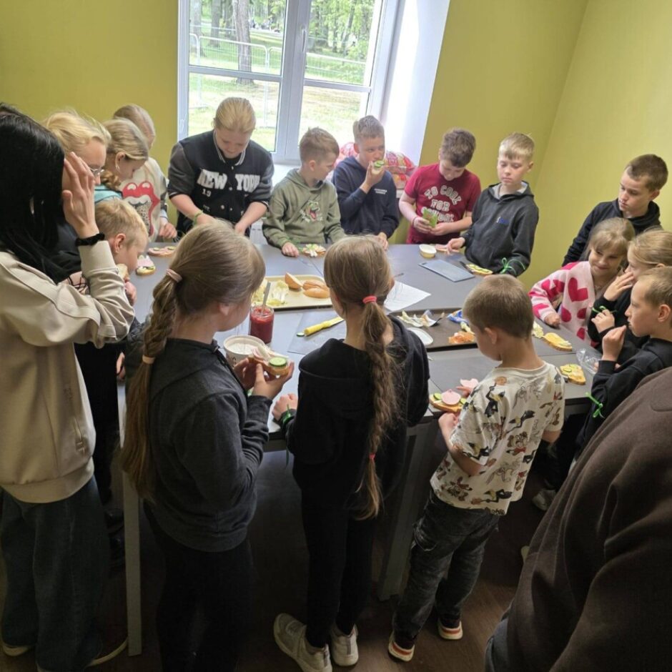 Bērnu un pieaugušo grupa sapulcējusies ap galdiem, gatavo un ēd atvērtās sviestmaizes. Meitene melnā sporta kreklā ar uzrakstu 