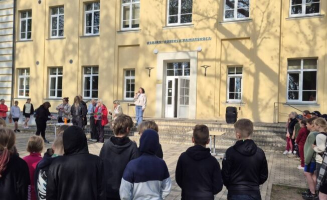 Cilvēku grupa, tostarp daudzi skolēni, pulcējusies pie gaiši dzeltenas skolas ēkas ar uzrakstu 