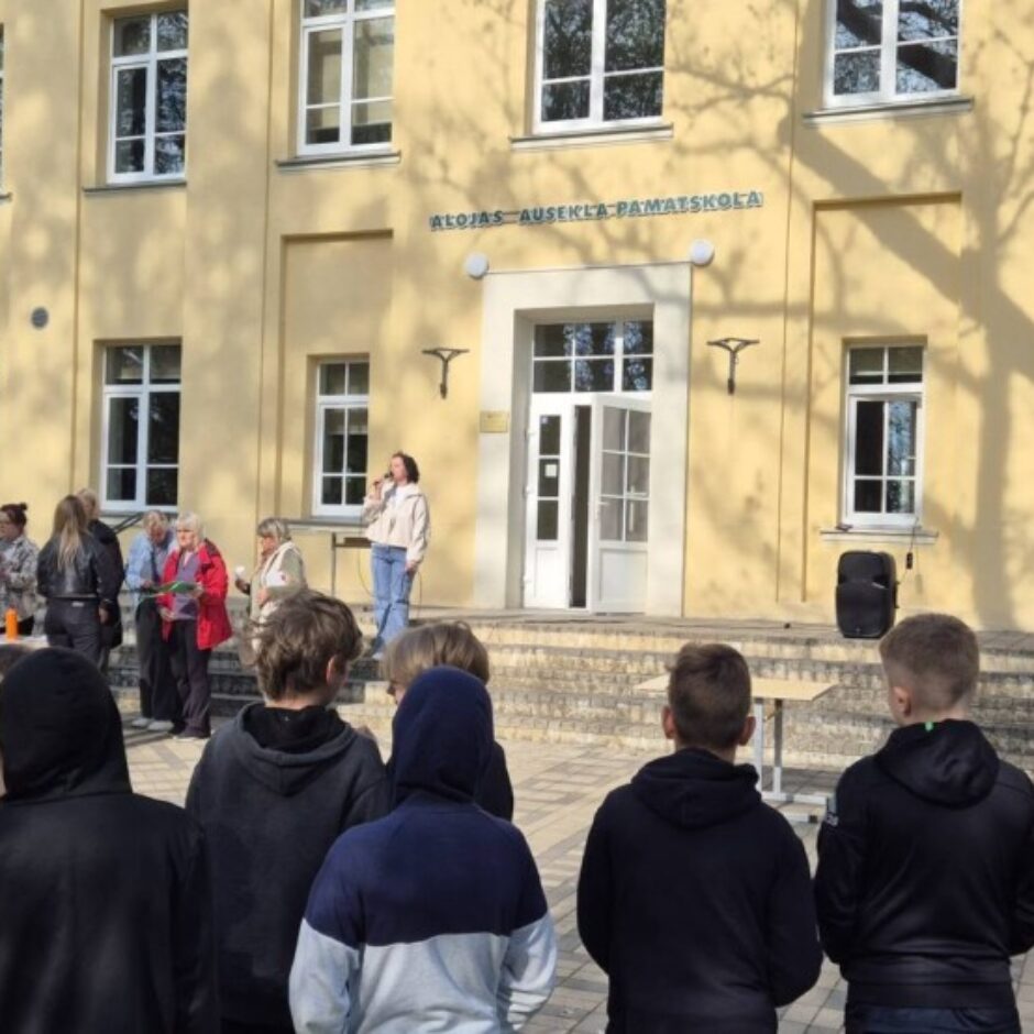 Cilvēku grupa, tostarp daudzi skolēni, pulcējusies pie gaiši dzeltenas skolas ēkas ar uzrakstu 
