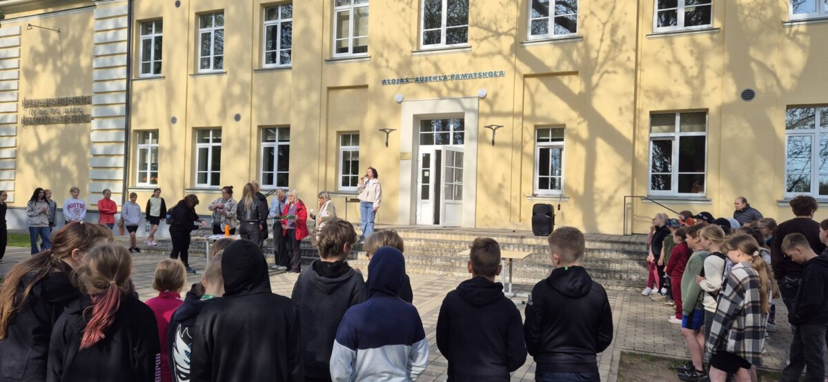 Cilvēku grupa, tostarp daudzi skolēni, pulcējusies pie gaiši dzeltenas skolas ēkas ar uzrakstu 