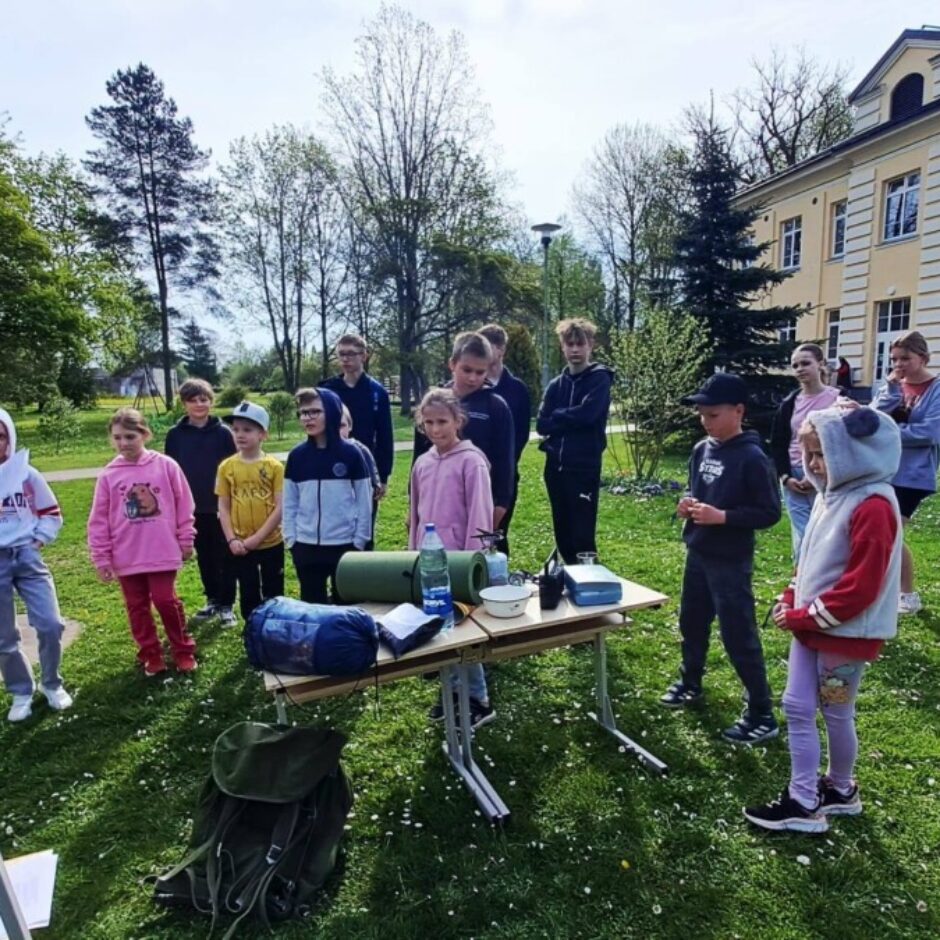 Pieaugusi sieviete instruē bērnu grupu zālājā pie galda ar kempinga aprīkojumu – guļammatraci, ūdens pudeli un gāzes plītiņu. Fonā redzama liela ēka.