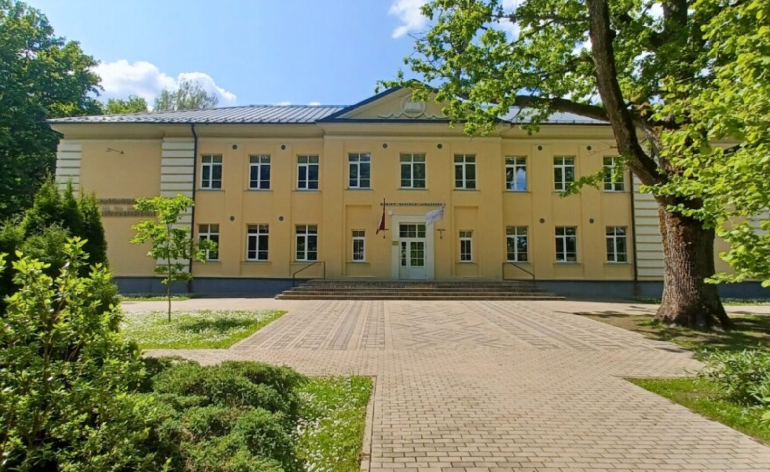 Dzeltena divstāvu ēka ar daudziem logiem un centrālo ieeju, pie kuras plīvo Latvijas karogs. Virs ieejas lasāms uzraksts 