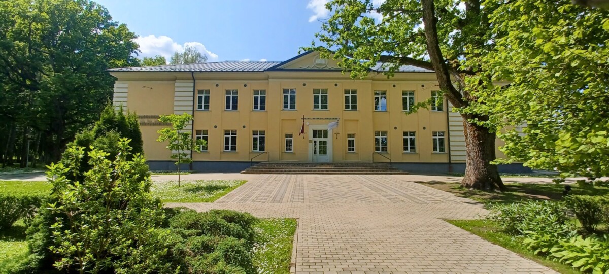 Dzeltena divstāvu ēka ar daudziem logiem un centrālo ieeju, pie kuras plīvo Latvijas karogs. Virs ieejas lasāms uzraksts 