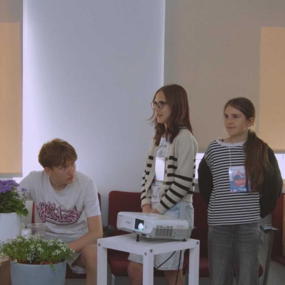 Trīs jaunieši gaišā telpā. Divas meitenes stāv pie balta projektora uz galda, abām ir kaklā identitātes kartes. Meitene centrā valkā brilles un svītrainu jaku. Pa labi stāvošā meitene ir svītrainā kreklā. Zēns sēž kreisajā pusē un skatās uz meitenēm. Priekšplānā ir telpaugi.