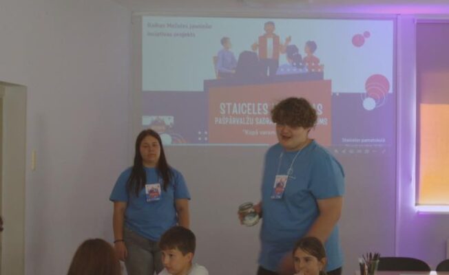 Jauniešu un bērnu grupa klases telpā ar projektora ekrānu fonā. Ekrānā redzamas ilustrācijas un teksts “Raidar Melnites jauniešu iniciatīvas projekts”, “STAICELES PASPARVALZU SADRA”, “Kopā varam” un “Staicele pamatskolā”. Jauns vīrietis zilā kreklā stāv un tur burku, savukārt citi jaunieši un bērni sēž pie galdiem.