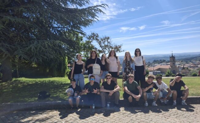 Grupa, kurā ir 13 jaunieši, no kuriem sešas meitenes stāv un septiņi zēni tup, pozē saulainā āra vidē. Aizmugurē pa kreisi ir liels zaļš koks, bet pa labi redzama pilsētas panorāma ar baznīcas torni un kalniem fonā. Uz vienas meitenes pleca somas ir teksts 