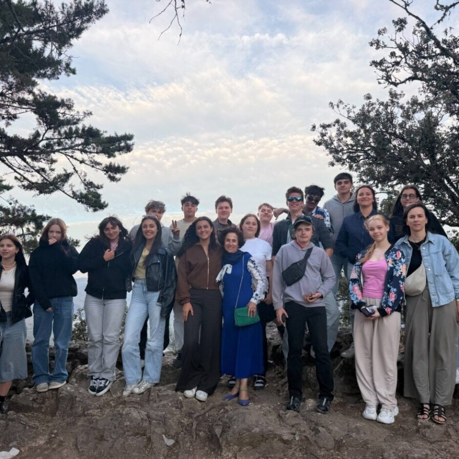 Jauniešu un pieaugušo grupa pozē fotogrāfijai uz akmeņainas kalna virsotnes ar kokiem un mākoņainām debesīm fonā.