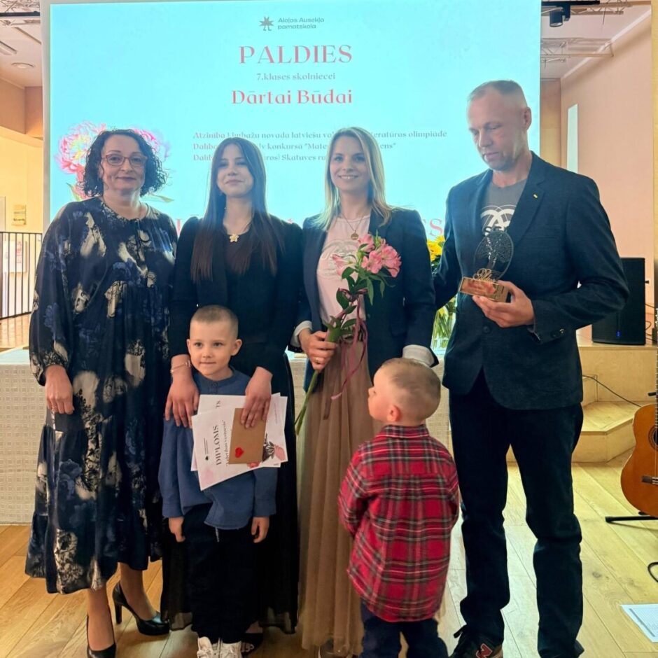 Seši cilvēki – trīs sievietes, divi mazi zēni un vīrietis – stāv grupā koka grīdas zālē pie ekrāna, uz kura rakstīts 