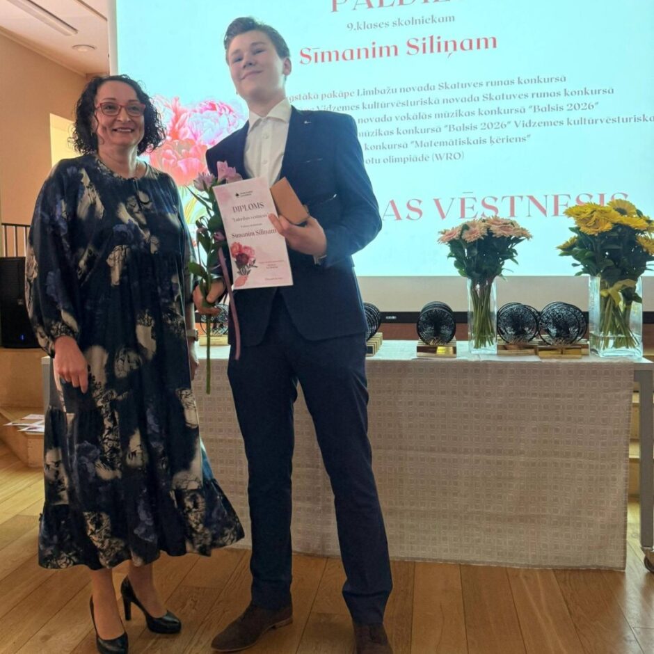Jauns vīrietis tumšā uzvalkā smaidot stāv blakus smaidīgai sievietei, rokās turot diplomu ar uzrakstu 