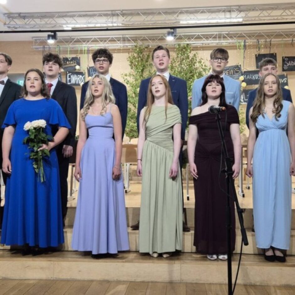 Vienpadsmit jauniešu grupa, seši puiši uzvalkos un piecas meitenes garās kleitās, stāv uz skatuves ar vairākiem pakāpieniem un dzied. Viena meitene tur baltu rožu pušķi. Fona dekorācijās redzami uzraksti 