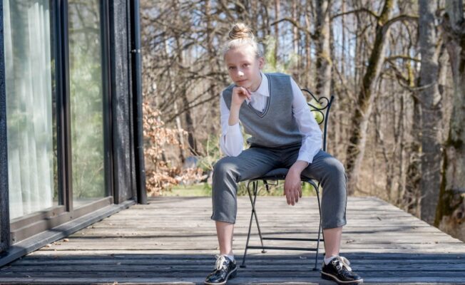 Jauna meitene skolas uniformā – baltā kreklā, pelēkā vestē un biksēs – sēž uz krēsla uz koka terases, ar roku uz zoda un skatās tieši kamerā. Fonā redzami koki un moderna ēka.