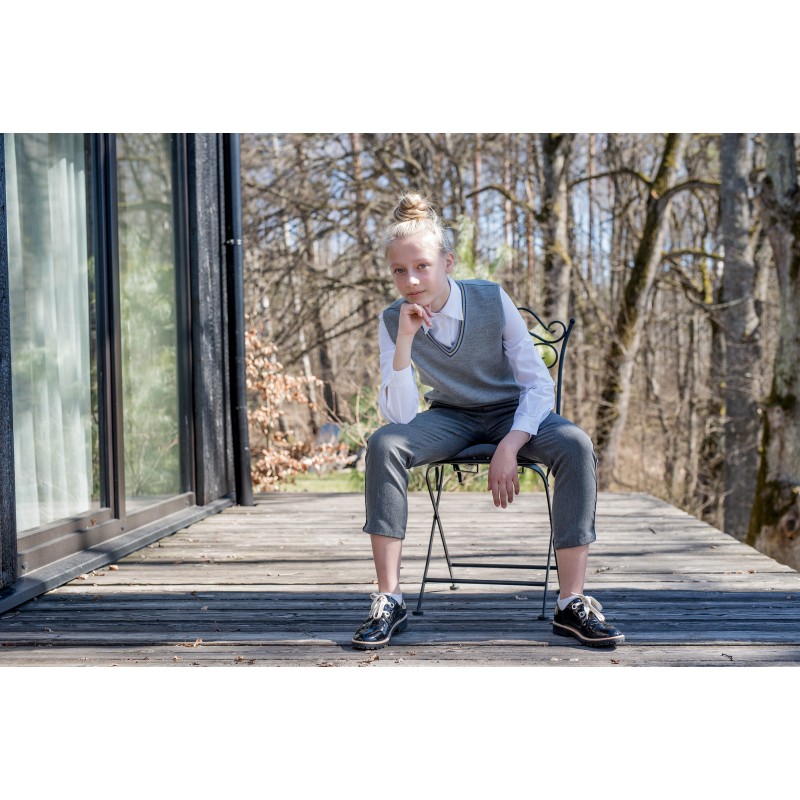 Jauna meitene skolas uniformā – baltā kreklā, pelēkā vestē un biksēs – sēž uz krēsla uz koka terases, ar roku uz zoda un skatās tieši kamerā. Fonā redzami koki un moderna ēka.