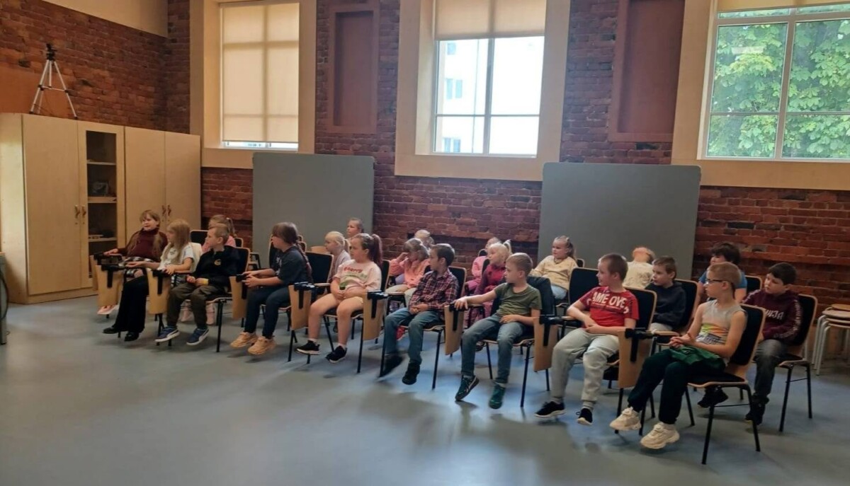 Bērnu grupa sēž krēslos telpā ar ķieģeļu sienām un lieliem logiem, uzmanīgi skatoties uz priekšu.