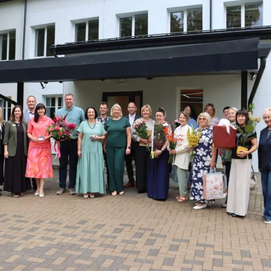 Liela grupa cilvēku, tostarp vīrieši un sievietes, smaidot pozē fotogrāfijai ārā pie baltas ēkas. Daudzi no viņiem tur ziedu pušķus un dāvanas.