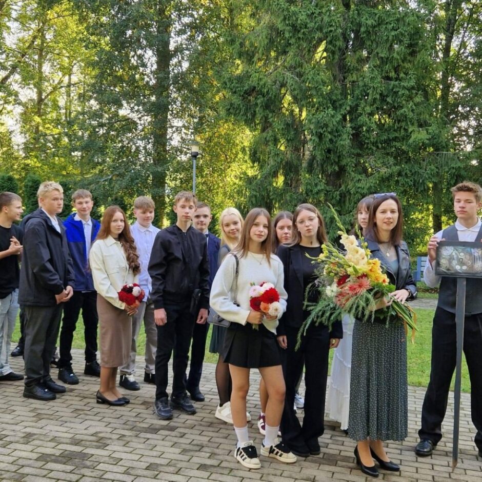 Studentu grupa, zēni un meitenes, stāv ārā uz bruģēta laukuma. Daudzi tur ziedus; viena sieviete tur lielu ziedu pušķi. Zēns pašā kreisajā pusē tur lāpstu, un zēns pašā labajā pusē tur ierāmētu attēlu uz kāta. Fonā redzami zaļi koki.