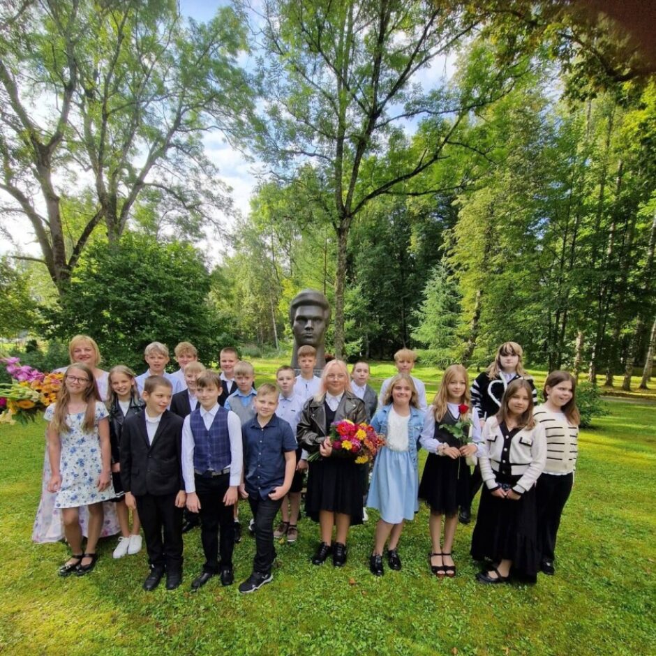 Bērnu un pieaugušo grupa pozē zaļā parkā ar kokiem fonā un lielu vīrieša krūšutēlu centrā. Daži grupas dalībnieki tur ziedus.