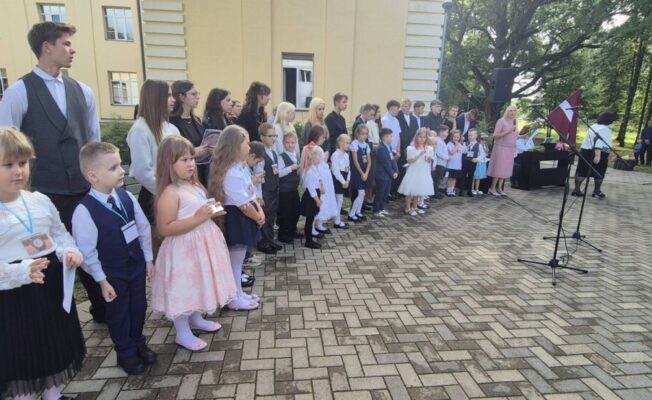 Skolēni un pieaugušie pulcējušies ārā pie skolas ēkas svinīgā pasākumā. Priekšplānā stāv mazi bērni, aiz tiem vecāki skolēni. Labajā pusē sieviete rozā kleitā runā mikrofonā, blakus redzams Latvijas karogs.
