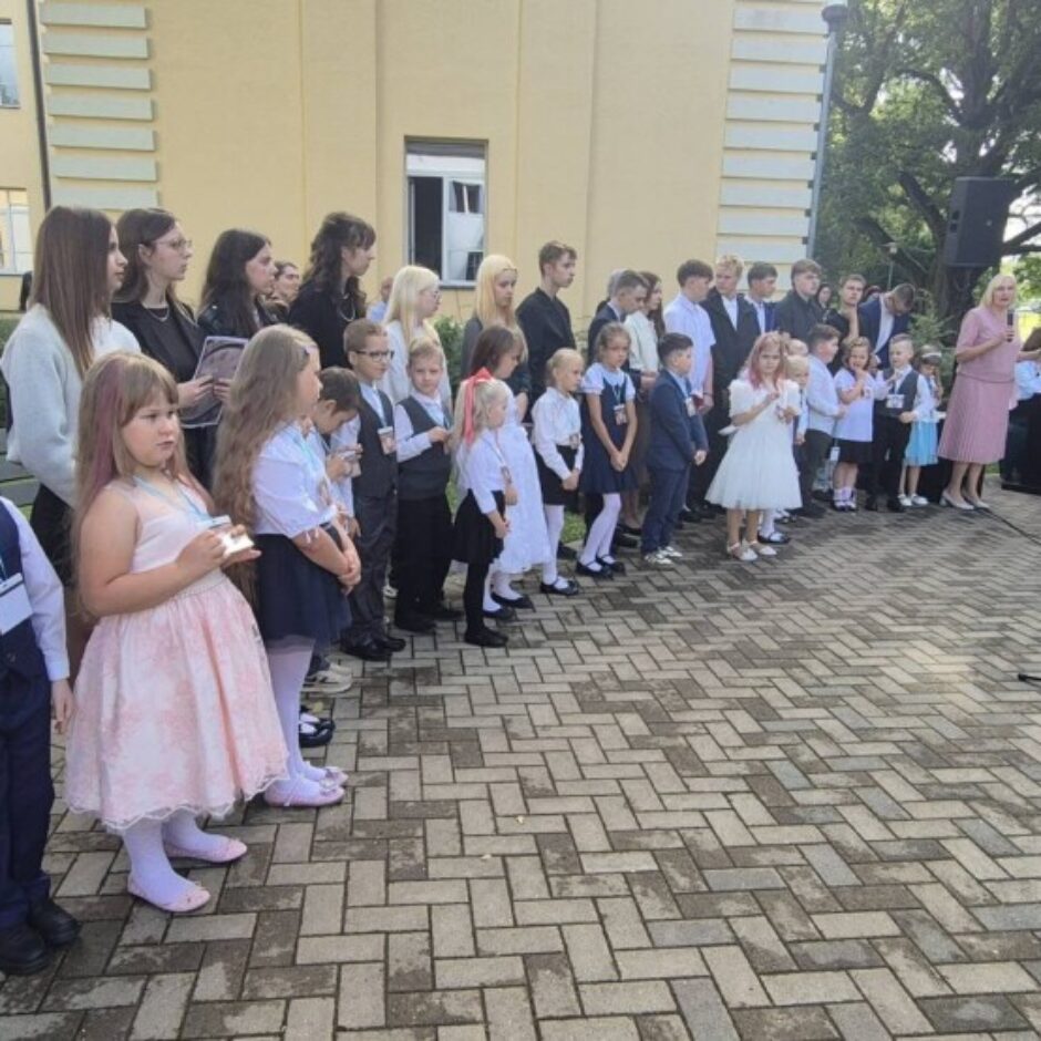 Skolēni un pieaugušie pulcējušies ārā pie skolas ēkas svinīgā pasākumā. Priekšplānā stāv mazi bērni, aiz tiem vecāki skolēni. Labajā pusē sieviete rozā kleitā runā mikrofonā, blakus redzams Latvijas karogs.