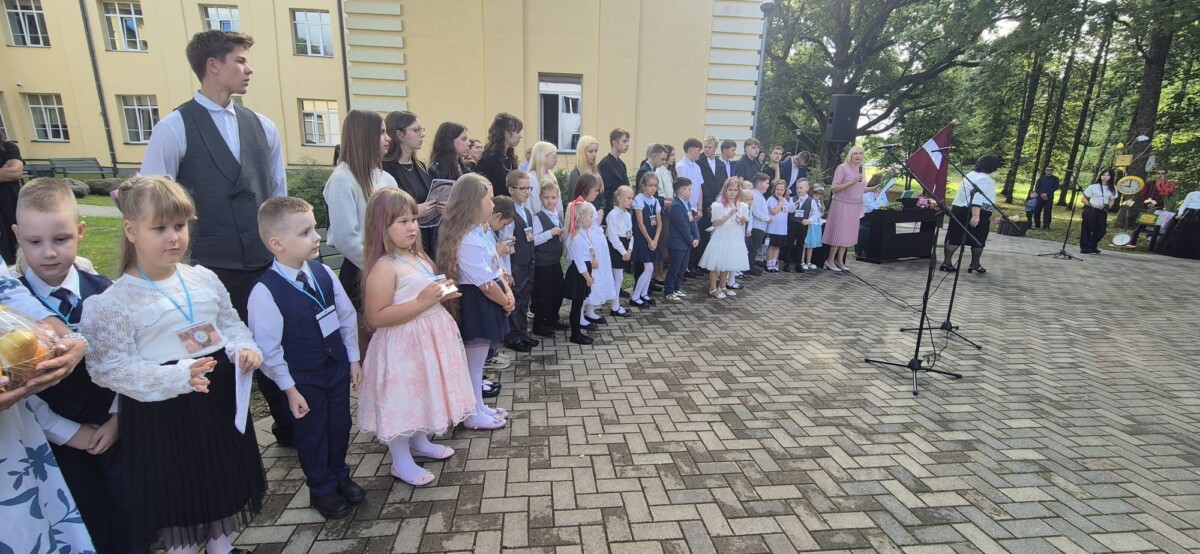 Skolēni un pieaugušie pulcējušies ārā pie skolas ēkas svinīgā pasākumā. Priekšplānā stāv mazi bērni, aiz tiem vecāki skolēni. Labajā pusē sieviete rozā kleitā runā mikrofonā, blakus redzams Latvijas karogs.
