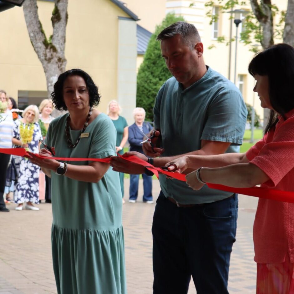 Divas sievietes un vīrietis griež sarkanu lentu ar šķērēm atklāšanas ceremonijā. Aiz viņiem stāv cilvēku grupa, un fonā redzama gaiša ēka un koki.