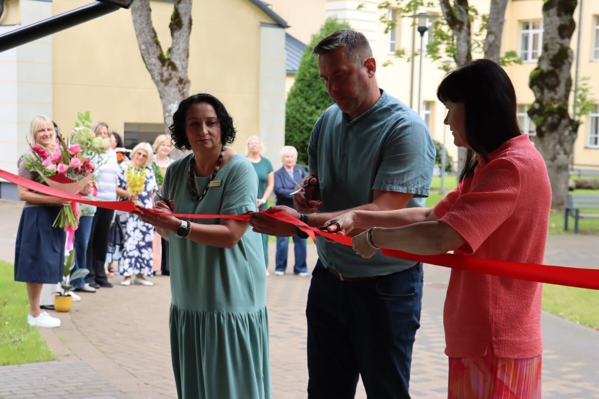 Divas sievietes un vīrietis griež sarkanu lentu ar šķērēm atklāšanas ceremonijā. Aiz viņiem stāv cilvēku grupa, un fonā redzama gaiša ēka un koki.