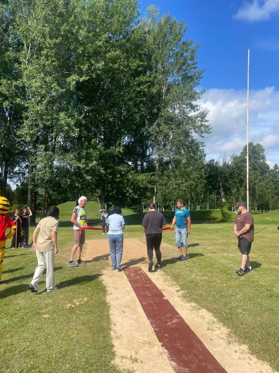 Vairāki pieaugušie pulcējas ap tāllēkšanas bedri ar sarkanbrūnu skrejceļu un smilšu nosēšanās zonu zaļā zālājā, ko ieskauj koki. Divi cilvēki tur sarkanu lentu pāri bedrei. Kreisajā pusē redzams cilvēks dzelteni sarkanā kostīmā. Fons ir zilas debesis ar dažiem mākoņiem.