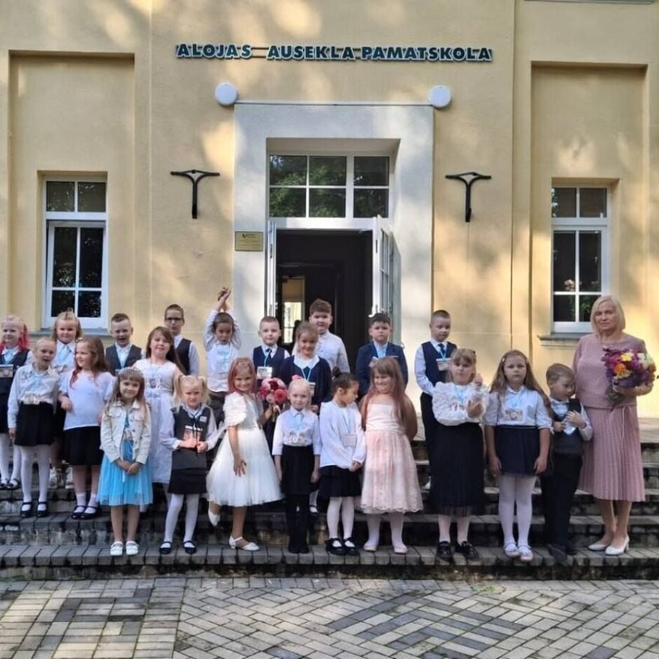 Grupa bērnu un sieviete ar ziedu pušķi stāv uz skolas 