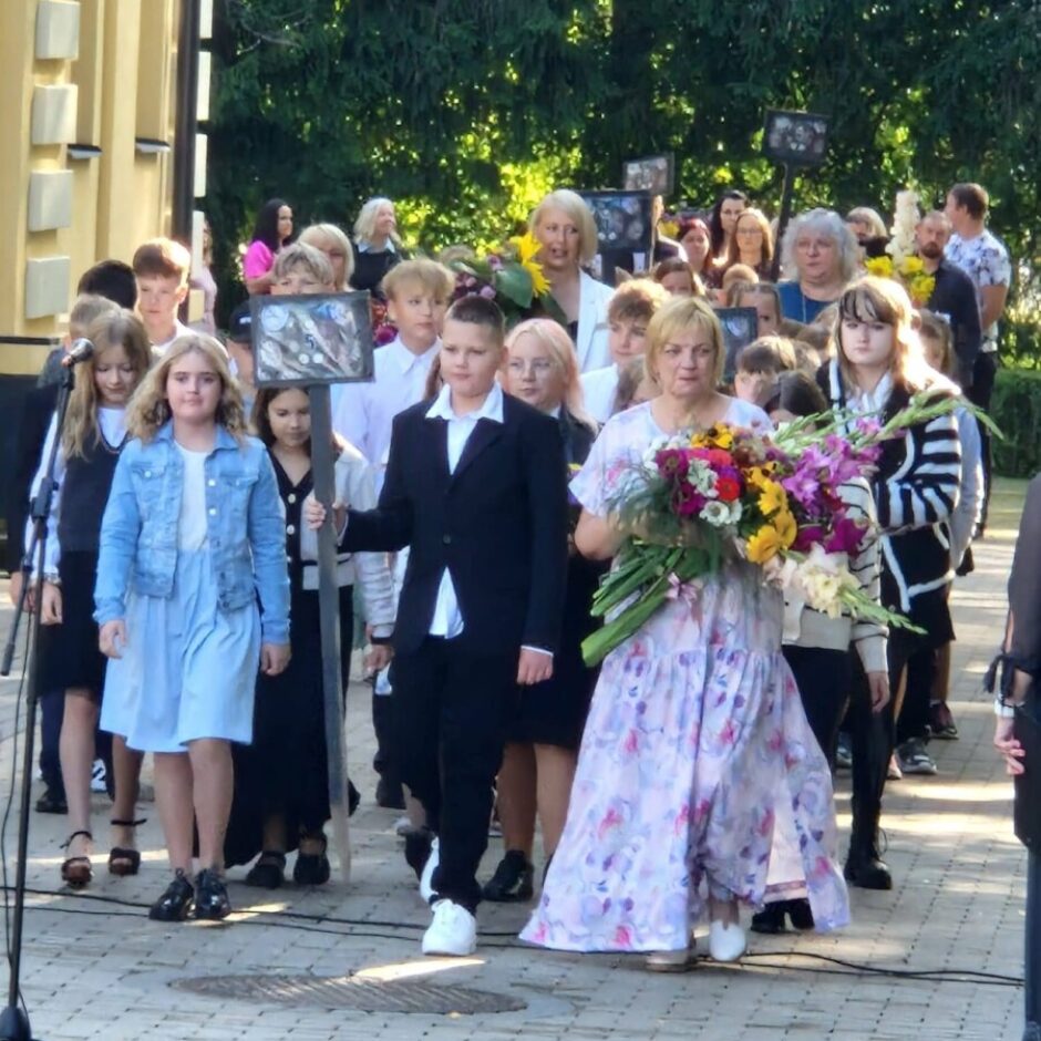 Skolēnu un pieaugušo gājiens brīvā dabā, priekšplānā sieviete garā ziedu kleitā nes lielu, krāsainu ziedu pušķi, bet zēns tumšā uzvalkā tur ierāmētu portretu uz kāta.