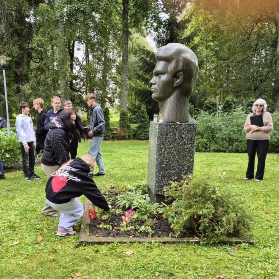 Jauniešu grupa un sieviete stāv ap jauna vīrieša granīta krūšutēlu uz pjedestāla parkā. Viens jaunietis liek ziedus pieminekļa pakājē, kurā aug arī citi ziedi un ir iedegtas sveces.
