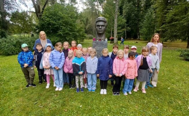 Grupa bērnu un divas sievietes pozē fotogrāfijai, stāvot uz zāles pie pieminekļa ar vīrieša krūšutēlu parkā.