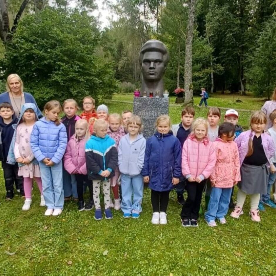 Grupa bērnu un divas sievietes pozē fotogrāfijai, stāvot uz zāles pie pieminekļa ar vīrieša krūšutēlu parkā.