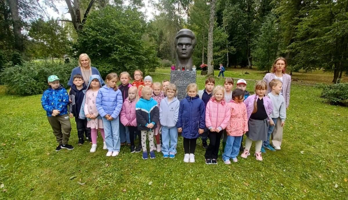 Grupa bērnu un divas sievietes pozē fotogrāfijai, stāvot uz zāles pie pieminekļa ar vīrieša krūšutēlu parkā.