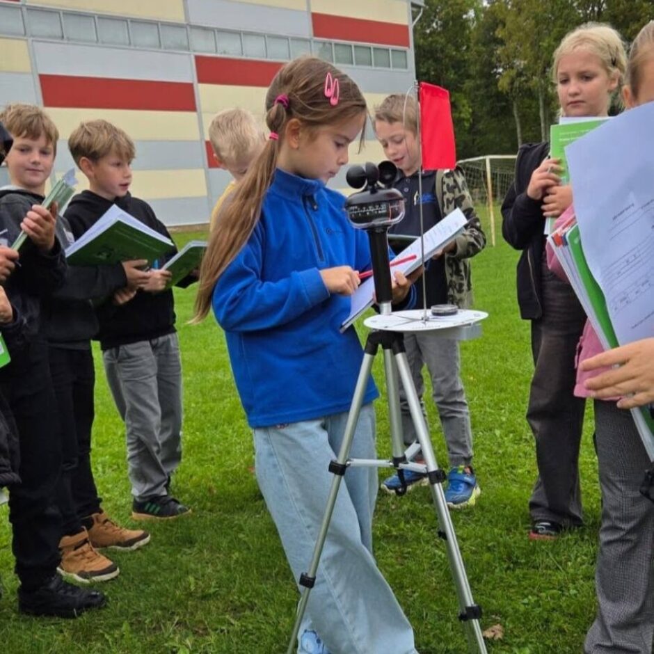 Bērnu grupa uz zālāja piedalās āra nodarbībā. Meitene zilā jakā veic mērījumus ar portatīvo meteoroloģisko staciju uz statīva, pierakstot datus. Apkārtējie bērni vēro ar grāmatiņām rokās. Fonā skolas ēka.