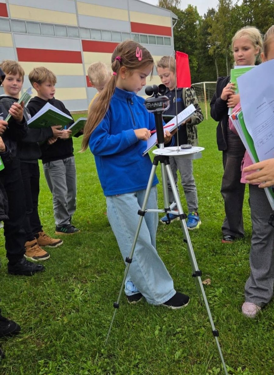 Bērnu grupa uz zālāja piedalās āra nodarbībā. Meitene zilā jakā veic mērījumus ar portatīvo meteoroloģisko staciju uz statīva, pierakstot datus. Apkārtējie bērni vēro ar grāmatiņām rokās. Fonā skolas ēka.