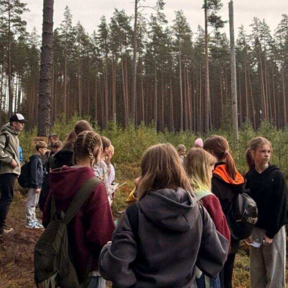 Skolēnu grupa stāv mežā, klausoties gidu ar lāpstu, kurš tērpies zaļā vestē. Apkārt redzamas augstas priedes un zems veģetācijas segums. Fonā uz zīmes lasāms vārds 