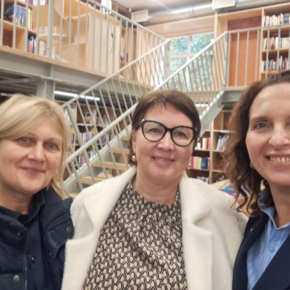 Trīs smaidošas sievietes pozē fotogrāfijai bibliotēkā vai grāmatnīcā ar grāmatu plauktiem un kāpnēm fonā.