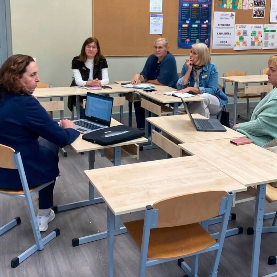Piecas pieaugušas sievietes sēž pie skolā izvietotiem galdiem, kas veido kopīgu sanāksmes vai apmācību telpu. Divām sievietēm ir atvērti portatīvie datori. Uz tāfeles fonā redzami vārdi: ATBILDĪBA, SADARBĪBA, CIENA.