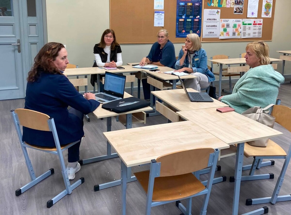 Piecas pieaugušas sievietes sēž pie skolā izvietotiem galdiem, kas veido kopīgu sanāksmes vai apmācību telpu. Divām sievietēm ir atvērti portatīvie datori. Uz tāfeles fonā redzami vārdi: ATBILDĪBA, SADARBĪBA, CIENA.