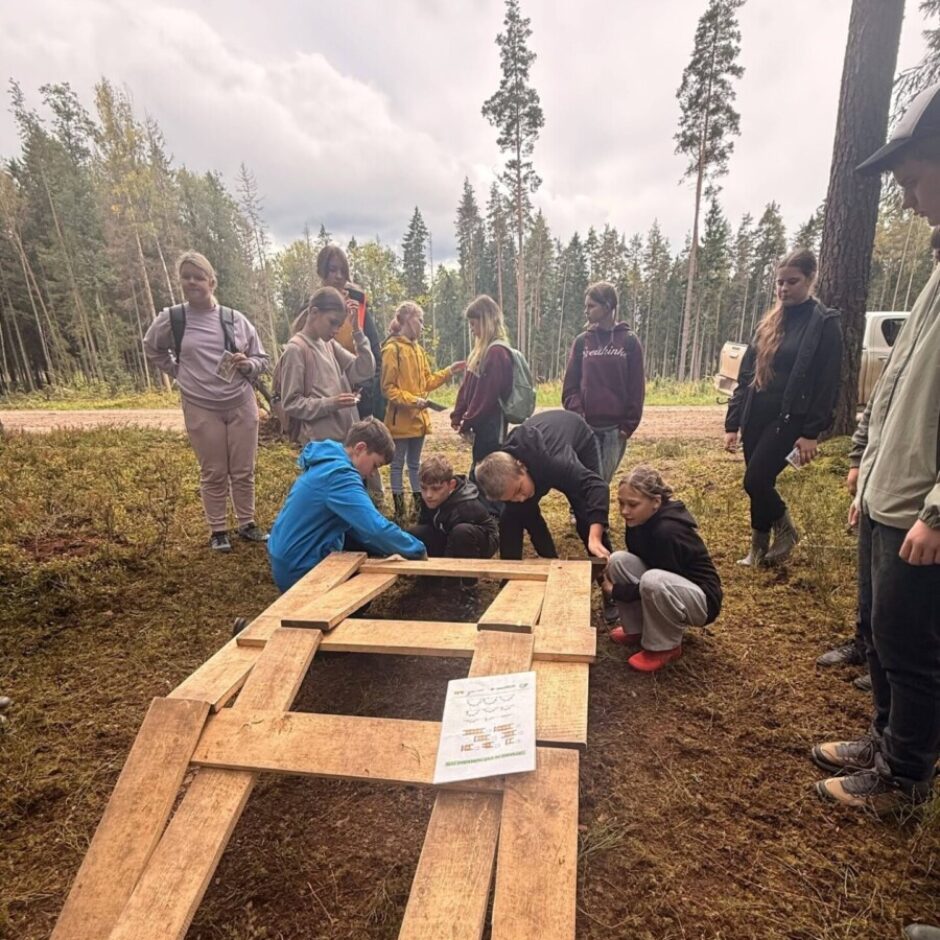 Mākoņainā dienā meža ielokā jaunieši un pieaugušie pulcējas ap uz zemes novietotu koka rāmi. Daži strādā pie tā, kamēr citi vēro. Uz rāmja ir lapa ar instrukcijām un simboliem.